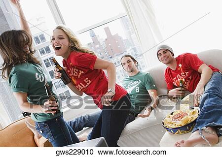 Stock Image - Two young men lying on a couch and two young women drinking beer and dancing. Fotosearch