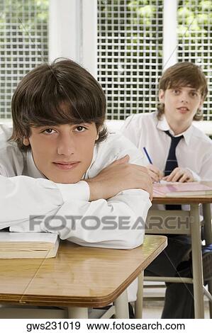 Portrait of a young man leaning on a desk with a teenage boy sitting behind him in a classroom View Large Photo Image Stock Photo - Portrait of a young man leaning on a desk with a teenage boy sitting behind him in a classroom. Fotosearch