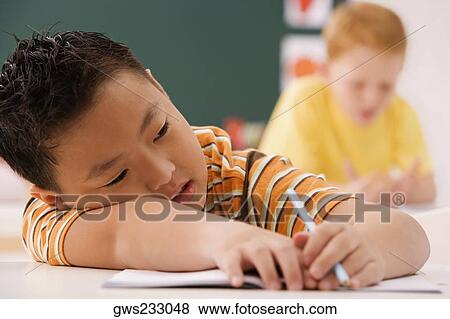 Stock Photo - Close-up of a schoolboy resting on a desk with another schoolboy sitting in the background in a classroom. Fotosearch