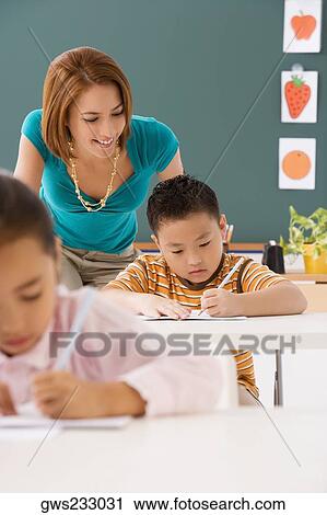 Close-up of a schoolboy writing on a notebook with a pencil with his female teacher standing behind him View Large Photo Image Stock Image - Close-up of a schoolboy writing on a notebook with a pencil with his female teacher standing behind him. Fotosearch