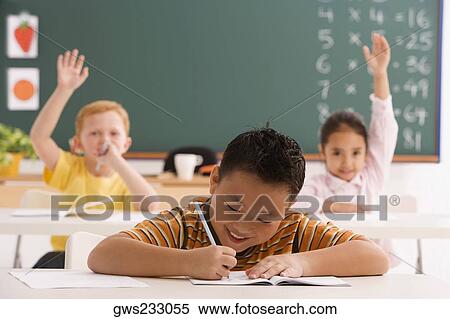 Schoolboy writing on a notebook with a pencil and two students sitting in the background with their hands raised View Large Photo Image Stock Photography - Schoolboy writing on a notebook with a pencil and two students sitting in the background with their hands raised. Fotosearch