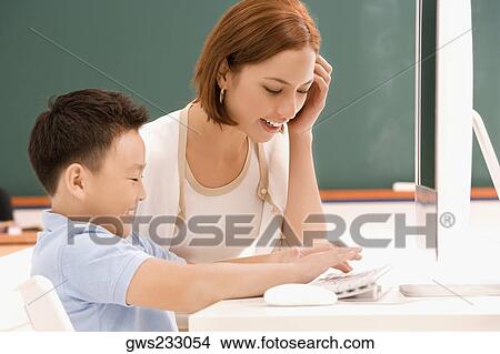 Picture - Side profile of a schoolboy using a computer with his female teacher sitting beside him in a classroom. Fotosearch