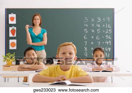 Three students sitting in a classroom with their teacher standing in the background View Large Photo Image Stock Image - Three students sitting in a classroom with their teacher standing in the background. Fotosearch