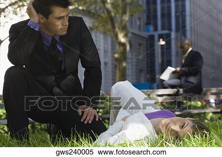 Stock Photography - Portrait of a young woman lying on grass in a park with a businessman beside her. Fotosearch