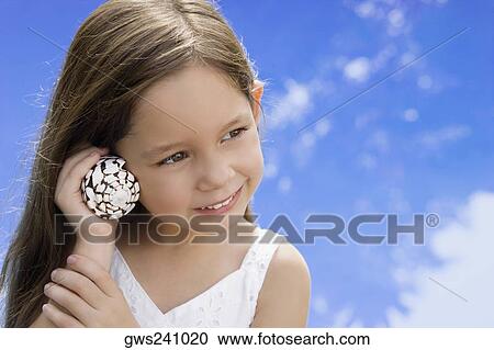 Stock Image - Close-up of a girl listening to shell and smiling. Fotosearch
