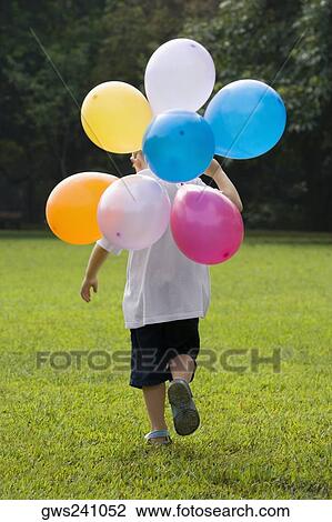 Stock Image - Rear view of a boy holding balloons and running in a playground. Fotosearch