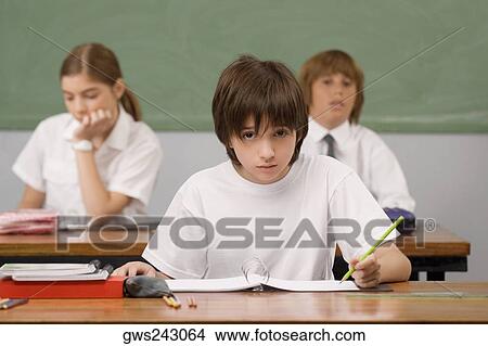 Schoolboy sitting at a desk with other students in the background View Large Photo Image Picture - Schoolboy sitting at a desk with other students in the background. Fotosearch