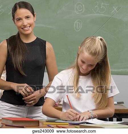 Stock Photo - Schoolgirl writing on a textbook with her female teacher standing behind her and smiling. Fotosearch