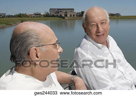Stock Image - Two senior men sitting on a bench at the lakeside. Fotosearch