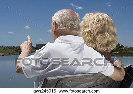 Stock Photograph - Rear view of a senior couple sitting on a bench at the lakeside. Fotosearch