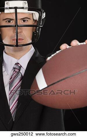 Stock Image - Portrait of a businessman wearing a football helmet and holding an American football. Fotosearch