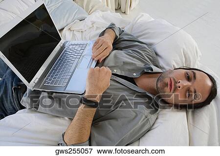 Stock Photo - High angle view of a young man lying on the bed and a laptop on his stomach. Fotosearch