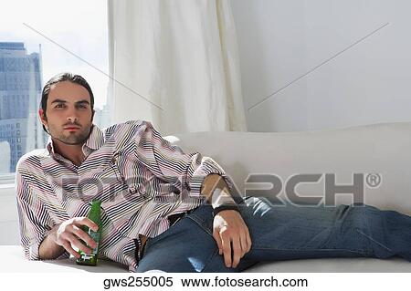Stock Photography - Portrait of a young man lying on a couch and holding a beer bottle. Fotosearch