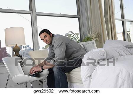 Stock Image - Portrait of a young man sitting on the bed and working on a laptop. Fotosearch