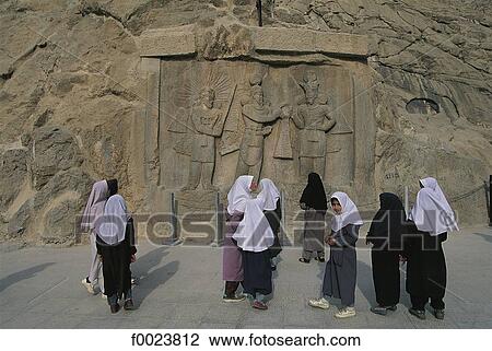 Stock Image - Iran, Kermanshah, Taq-e Bostan, veiled women in front of high relief of crowning ceremony of Ardashir. Fotosearch