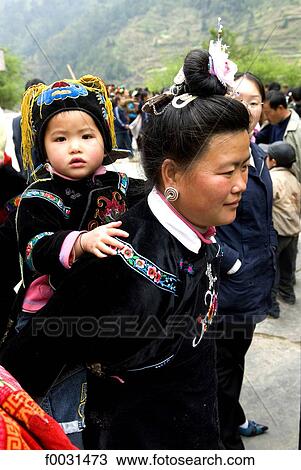Stock Image - China, Guizhou, Taojiang village, Miao woman in traditional costume carrying her child on her back. Fotosearch