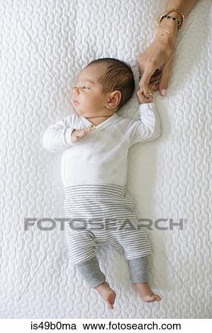 Stock Image - Baby boy lying on bed, mother holding baby's hand, overhead view. Fotosearch