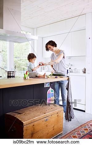 Baby girl sitting on kitchen counter watching mother slicing cucumber View Large Photo Image Picture - Baby girl sitting on kitchen counter watching mother slicing cucumber. Fotosearch