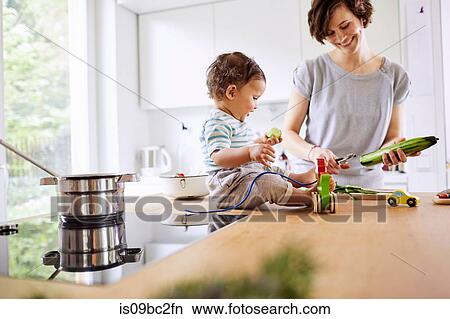 Picture - Baby girl sitting on kitchen counter watching mother slicing cucumber. Fotosearch