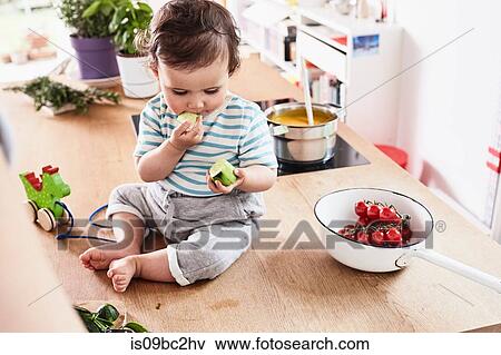 Stock Photo - Baby girl sitting on kitchen counter, eating cucumber. Fotosearch
