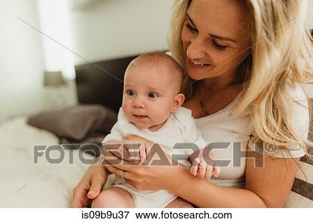 Stock Photo - Baby girl sitting on mother's lap on bed. Fotosearch