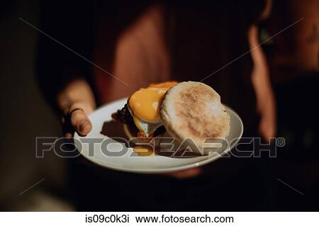 Stock Image - Barista holding plate with fried egg muffin in cafe, close up low key. Fotosearch