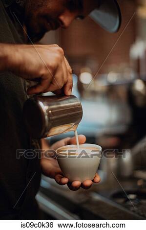 Stock Photograph - Barista pouring milk into cup of latte in cafe, close up of hands. Fotosearch