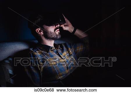 Bearded mid adult man sitting on sofa in darkness looking up through virtual reality headset View Large Photo Image Stock Photo - Bearded mid adult man sitting on sofa in darkness looking up through virtual reality headset. Fotosearch