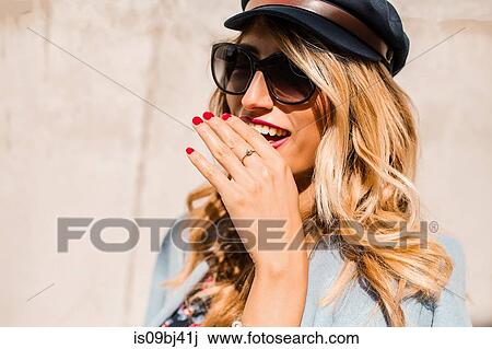 Stock Image - Blond, stylish mid adult woman wearing sunglasses in front of wall with hand covering mouth. Fotosearch