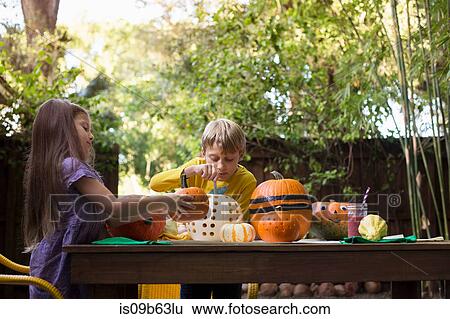 Stock Photograph - Boy and sister carving pumpkins on garden table. Fotosearch