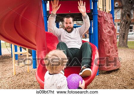 Boy looking at father playing on slide View Large Photo Image Stock Photograph - Boy looking at father playing on slide. Fotosearch