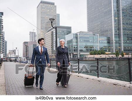 Stock Image - Businessman and businesswoman pulling trolley luggage, Canary Wharf, London, UK. Fotosearch