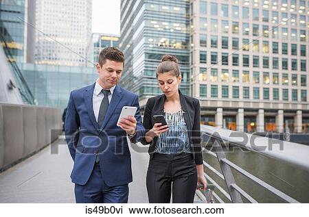 Stock Image - Businessman and businesswoman using mobile phone and pulling trolley luggage, Canary Wharf, London, UK. Fotosearch