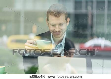 Stock Photograph - Businessman sitting in cafe, drinking coffee, using smartphone, viewed through window. Fotosearch