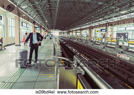 Businessman with wheeled suitcase, standing at train station, waiting for train View Large Photo Image Stock Image - Businessman with wheeled suitcase, standing at train station, waiting for train. Fotosearch