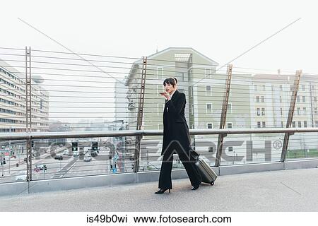 Stock Image - Businesswoman using mobile and pulling trolley luggage, Milan, Italy. Fotosearch