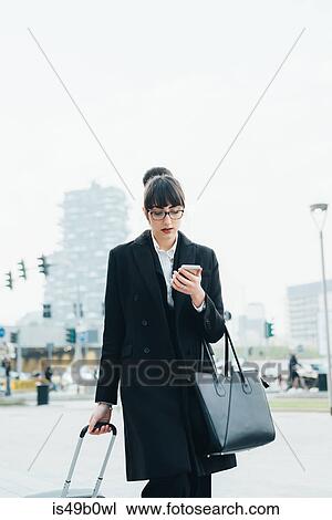 Stock Image - Businesswoman using mobile and pulling trolley luggage, Milan, Italy. Fotosearch