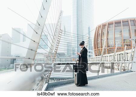 Stock Image - Businesswoman with trolley luggage on pedestrian bridge, Milan, Italy. Fotosearch