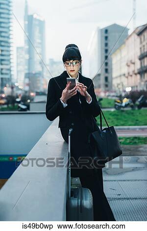 Stock Photography - Businesswoman with trolley luggage using mobile waiting by wall, Milan, Italy. Fotosearch