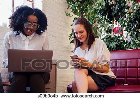 Stock Image - Colleagues sitting together on sofa in office using laptop. Fotosearch