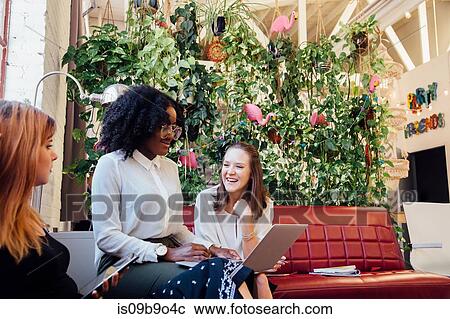 Stock Image - Colleagues sitting together on sofa in office using laptop. Fotosearch