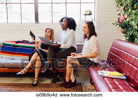 Stock Image - Colleagues sitting together on sofa in office using laptop. Fotosearch