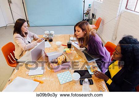 Stock Photo - Colleagues working together at desk sharing doughnuts. Fotosearch
