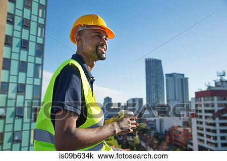Stock Image - Construction worker outdoors, holding takeaway coffee cup, elevated view of surrounding buildings. Fotosearch