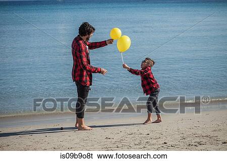 Stock Photograph - Father and son on beach, son holding two yellow balloons. Fotosearch