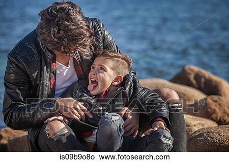 Father and son outdoors, fooling around, sitting on rocks beside sea View Large Photo Image Picture - Father and son outdoors, fooling around, sitting on rocks beside sea. Fotosearch
