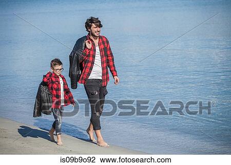Stock Image - Father and son running along beach, carrying jackets over shoulder. Fotosearch