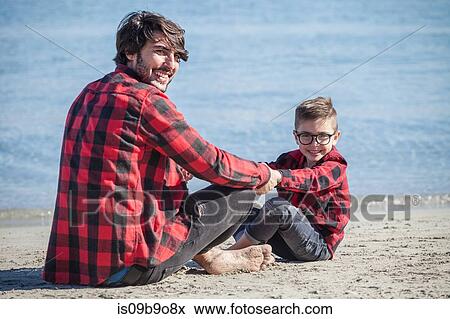 Father and son sitting face to face on beach, holding hands, smiling View Large Photo Image Stock Photo - Father and son sitting face to face on beach, holding hands, smiling. Fotosearch