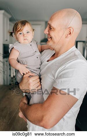 Stock Image - Father carrying baby daughter in living room, portrait. Fotosearch