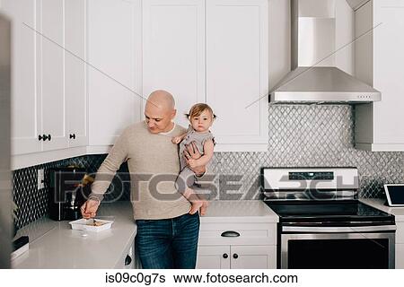 Stock Photograph - Father carrying baby daughter in kitchen, portrait. Fotosearch